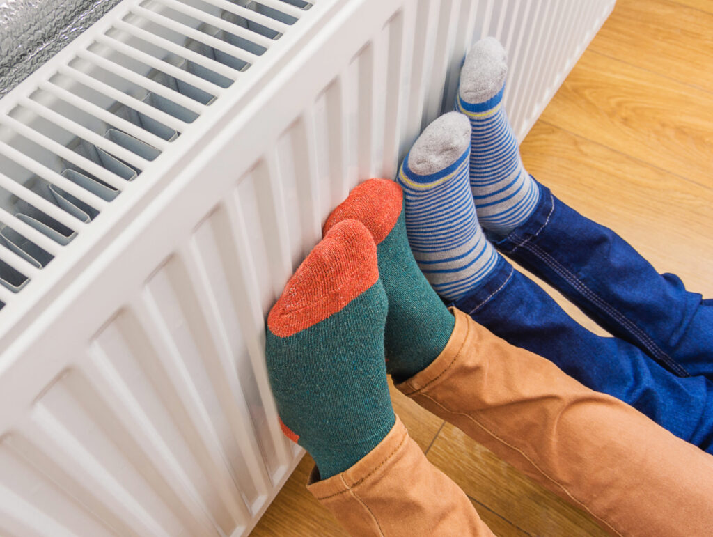 Woman and child wearing colorful pair of woolly socks warming cold feet in front of heating radiator in winter time. Electric or gas heater at home. Part of body, selective focus.